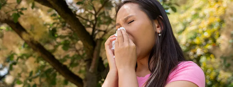 A person sneezing in a park, representing common symptoms of seasonal allergies.